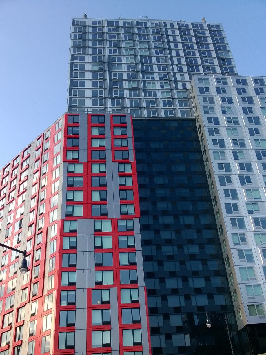 Looking up at 461 Dean Street in Brooklyn: a tall modular tower with red-framed, grey and dark glazed façade sections
