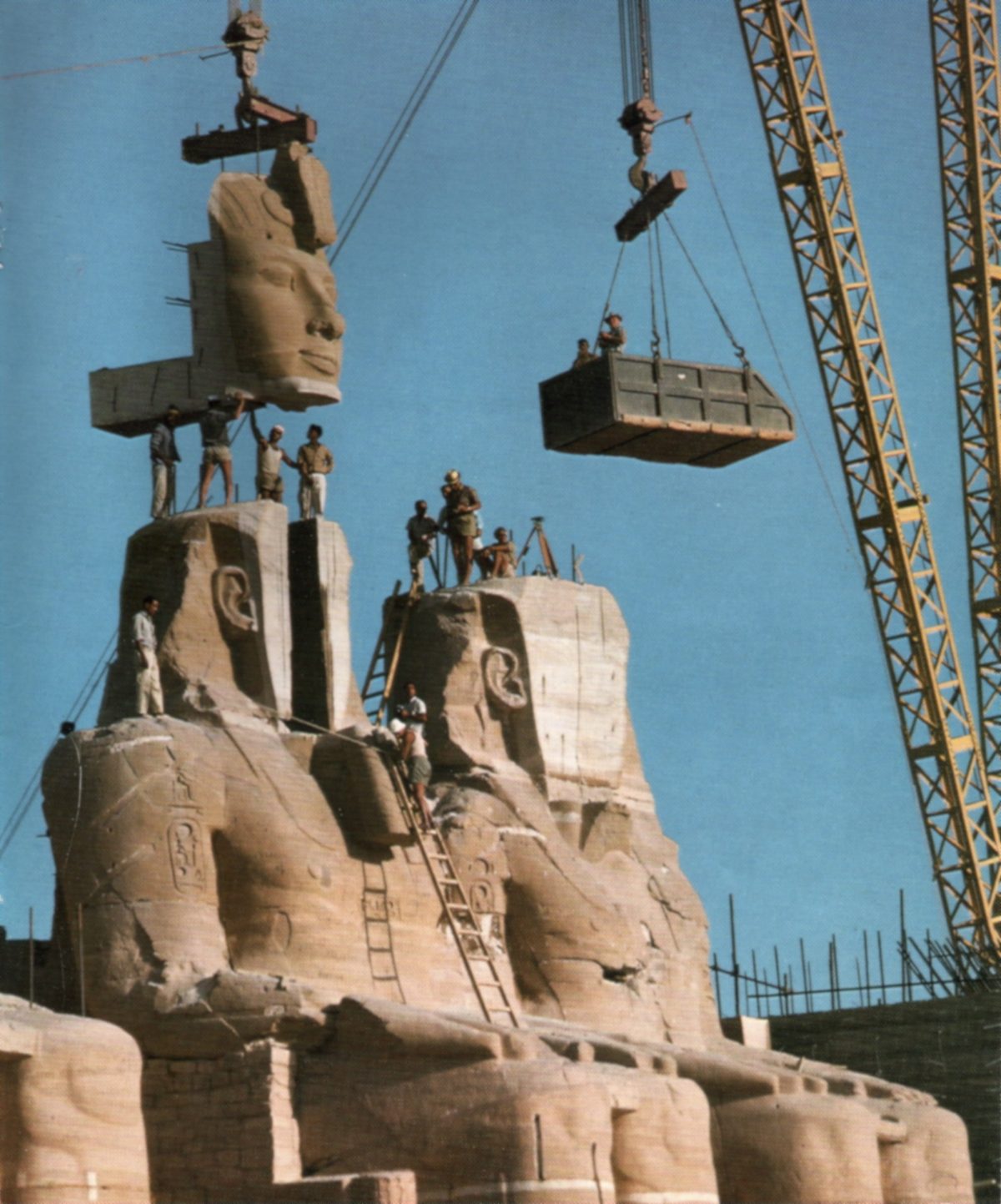 Workers on the reassembled colossal statues of Ramses II at the Great Temple of Abu Simbel as a crane lowers the statue face into place and a second crane holds a work platform