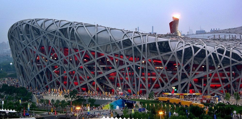The Beijing National Stadium at dusk during the 2008 Olympics, its interwoven steel facade lit red from inside and the Olympic cauldron burning on the roof
