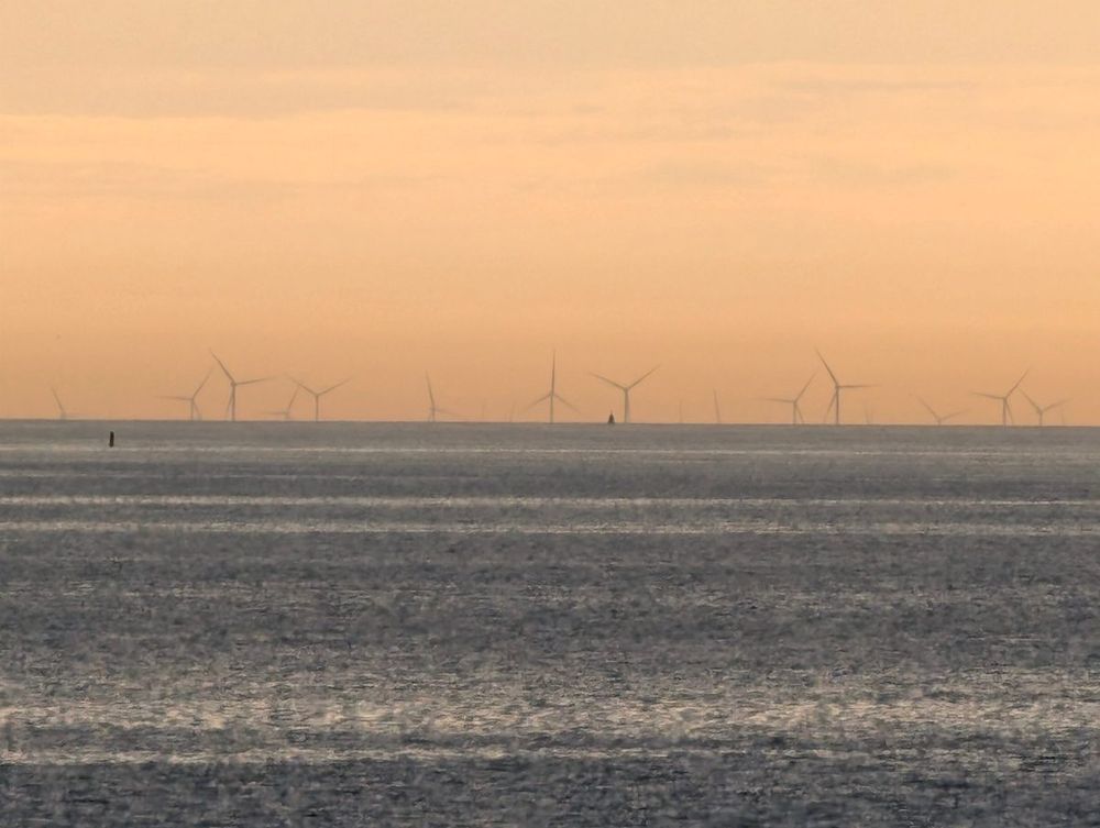 A row of offshore wind turbines on the horizon at sunset, seen across a calm grey sea under an orange sky