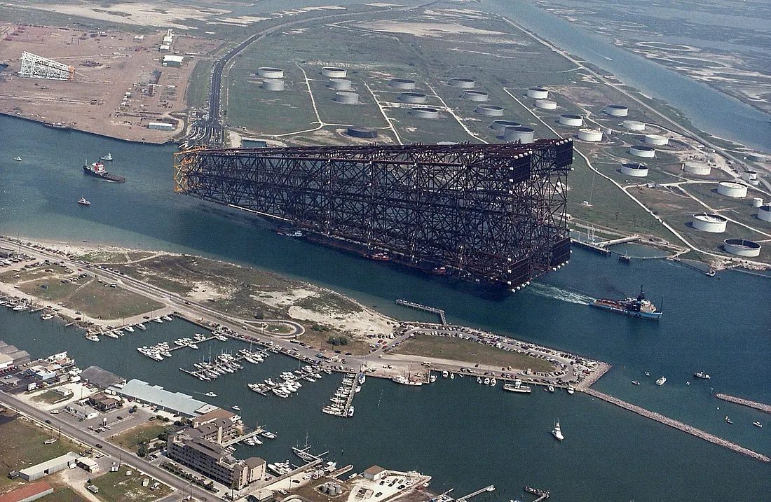 Aerial view of the Bullwinkle jacket under tow on its launch barge through the channel near Port Aransas, Texas, in 1988, dwarfing the tugs alongside