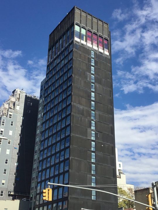 The citizenM New York Bowery hotel tower, a dark slender block with a grid of windows, seen from street level against a blue sky