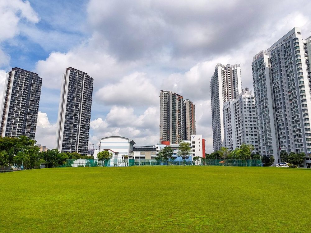 High-rise residential towers in Clementi, Singapore, including the Clement Canopy and Clavon condominiums, seen across a wide grass playing field under cloud