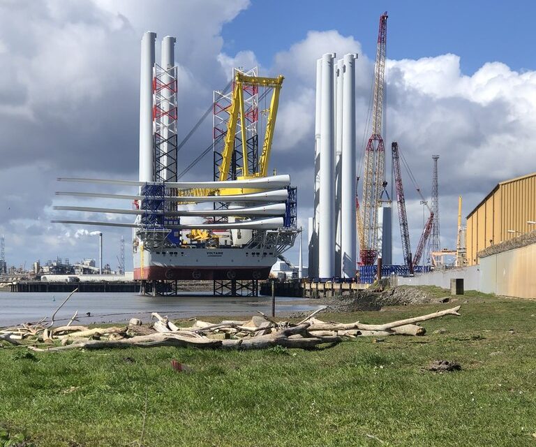 A jack-up installation vessel loaded with wind turbine blades and tower sections at a quayside, with further towers and crawler cranes behind