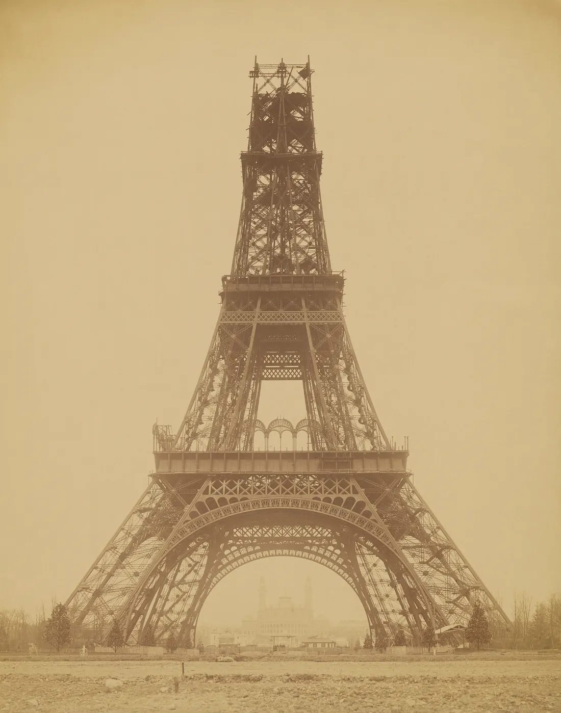 Sepia photograph of the Eiffel Tower under construction in 1888, the four inclined lattice legs risen past the first platform with timber scaffolding at the upper level