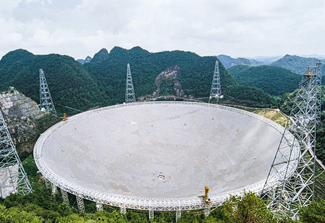 Aerial view of the FAST radio telescope's 500 metre spherical dish nested inside a green karst depression in Guizhou, ringed by white lattice support towers