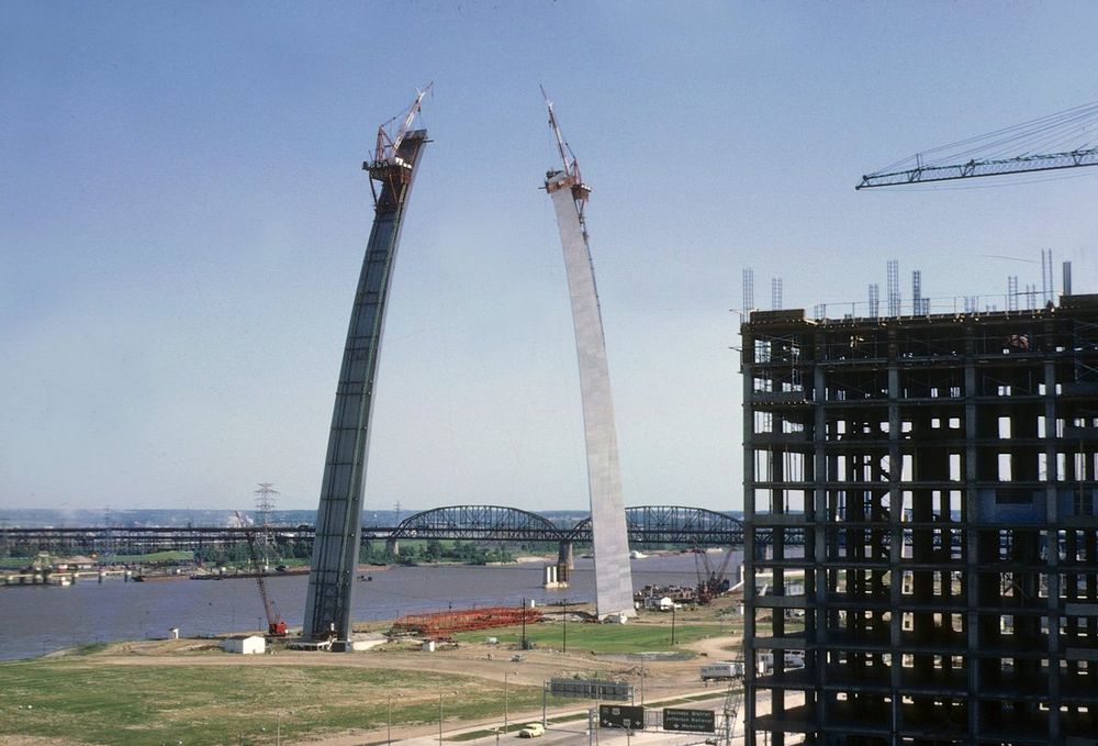 The two unfinished legs of the Gateway Arch rising towards each other in June 1965, each topped by a creeper crane, with the red steel truss lying on the ground between them and the Mississippi River behind