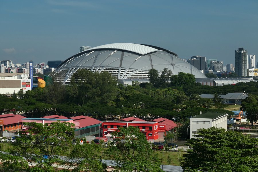 The Singapore Sports Hub's retractable dome roof viewed from a distance across trees and low buildings, its ribbed steel structure visible against the city skyline