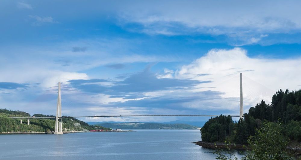 The Skarnsund cable-stayed bridge spanning a wide fjord strait between two tall concrete towers, with forested shores under a blue sky