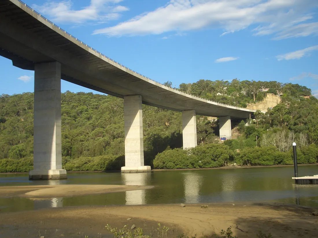 The Woronora River Bridge seen from the riverbank, its curved deck carried on tall tapered concrete piers above the water with bushland on both banks