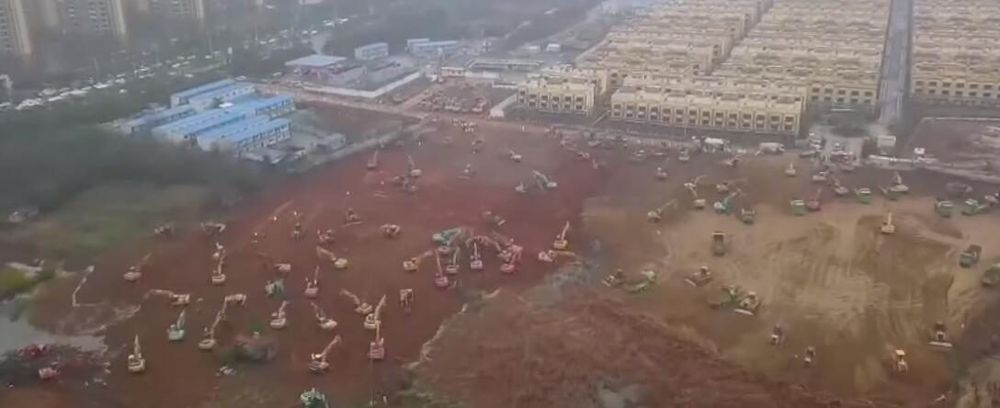 Aerial view of dozens of excavators and bulldozers working across a cleared red-earth site in Wuhan in January 2020, with residential blocks behind