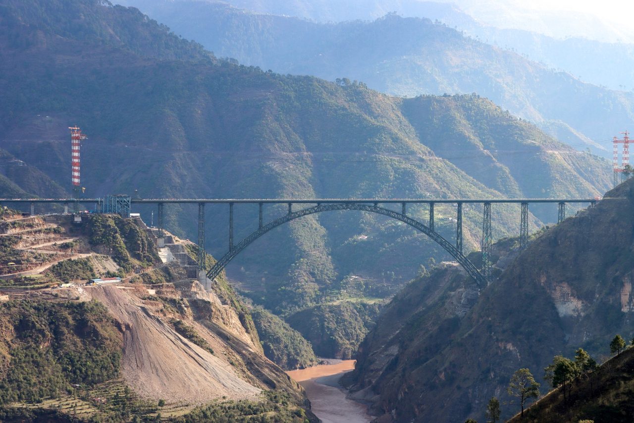 The completed Chenab Rail Bridge steel arch spanning the Chenab River gorge in Reasi district, Jammu and Kashmir, with the red-and-white cable-crane pylons still standing on both banks