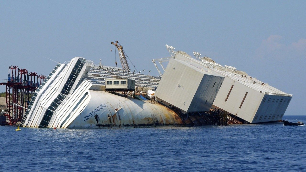 The capsized Costa Concordia lying on its side at Isola del Giglio with white buoyancy caissons fixed along the exposed hull ahead of the parbuckling operation