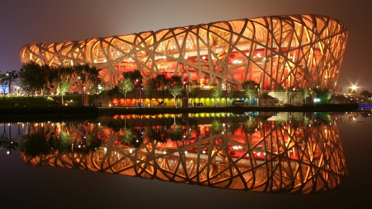 Beijing National Stadium at night, its interwoven steel lattice facade lit from within and reflected in still water