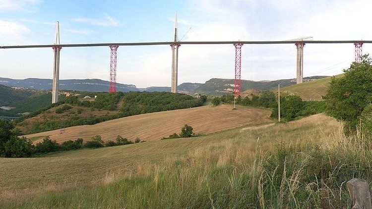 Millau Viaduct under construction with red temporary steel towers standing between the permanent concrete piers, supporting the launched deck