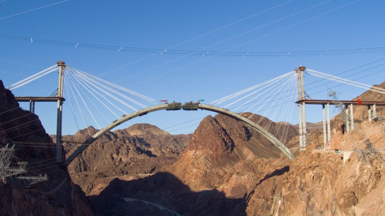 The Hoover Dam Bypass concrete arch under construction, both cantilevered halves reaching toward the crown, supported by temporary stay cables from twin towers